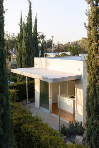 Los Angeles hillside ADU with smooth stucco walls, slim roofline, and sliding glass doors surrounded by lush greenery and tall Italian cypress trees.